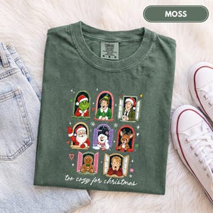 May include: A moss green t-shirt featuring a graphic print of various Christmas characters in individual window frames, including a green creature, an elf, a man, Santa Claus, a snowman, a reindeer, a gingerbread man, and a boy, with the text "too cozy for christmas" below.