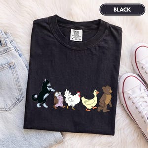 K&ouml;nnte beinhalten: Ein schwarzes T-Shirt mit einem Grafikdruck, der eine schwarz-wei&szlig;e Katze, eine braune Eule, ein wei&szlig;es Huhn, eine gelbe Ente und einen braunen B&auml;ren in einer Reihe zeigt. Das Shirt hat ein Comfort Colors Etikett am Hals und den Text BLACK.