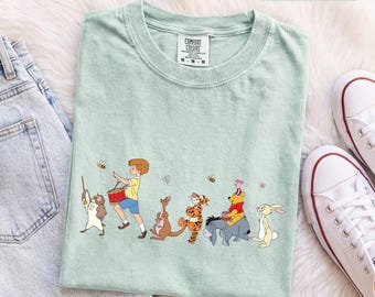 Könnte beinhalten: Ein hellgrünes T-Shirt mit einem Grafikdruck von Christopher Robin, der eine Trommel spielt, gefolgt von Kanga, Tigger, I-Aah mit Winnie Puuh auf dem Rücken und Rabbit. Das T-Shirt hat einen Rundhalsausschnitt und ein Comfort Colors Markenetikett.