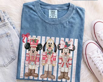 Könnte beinhalten: Ein blaues T-Shirt mit einer Grafik von Mickey Mouse, Pluto und Minnie Mouse als Nussknacker verkleidet. Die Figuren sind vor einem vertikal gestreiften Hintergrund platziert. Das Shirt hat einen Rundhalsausschnitt und ein Comfort Colors Etikett.