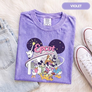 Könnte beinhalten: Ein violettes Kurzarm-T-Shirt mit einem Grafikdruck von Disney-Figuren wie Mickey Maus, Minnie Maus, Donald Duck, Daisy Duck, Goofy und Pluto vor der Epcot Spaceship Earth-Struktur mit dem aufgedruckten Schriftzug Epcot.