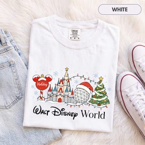 May include: A white short-sleeve t-shirt featuring a graphic of a castle, a geodesic dome wearing a Santa hat, a Christmas tree, and a Mickey Mouse ornament. The text Walt Disney World is printed below the graphic in a black script font.