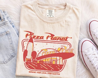 Könnte beinhalten: Ein elfenbeinfarbenes Kurzarm-T-Shirt mit einer roten und gelben Grafik eines Raketenschiffs und eines Gebäudes mit dem Text Pizza Planet. Die Grafik enthält den Schriftzug Serving Your Local Star Cluster. Das T-Shirt hat ein Comfort Colors Etikett.