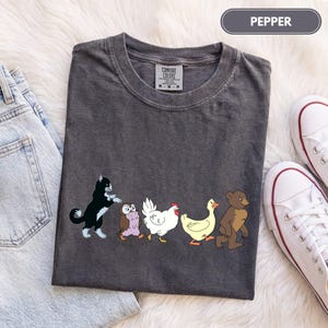 K&ouml;nnte beinhalten: Ein anthrazitfarbenes Kurzarm-T-Shirt mit einem Grafikdruck einer schwarz-wei&szlig;en Katze, einer braunen Eule, einem wei&szlig;en Huhn, einer gelben Ente und einem braunen B&auml;ren, die in einer Reihe laufen. Das Shirt hat ein Comfort Colors Etikett am Hals.