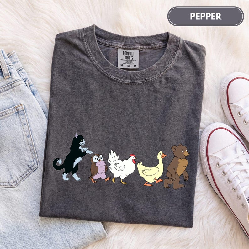 K&ouml;nnte beinhalten: Ein anthrazitfarbenes Kurzarm-T-Shirt mit einem Grafikdruck einer schwarz-wei&szlig;en Katze, einer braunen Eule, einem wei&szlig;en Huhn, einer gelben Ente und einem braunen B&auml;ren, die in einer Reihe laufen. Das Shirt hat ein Comfort Colors Etikett am Hals.