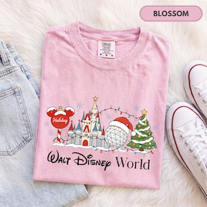 May include: A light pink short-sleeve t-shirt featuring a graphic of a holiday-themed castle, a geodesic sphere wearing a Santa hat, a Christmas tree, and a Mickey Mouse ornament. The text Walt Disney World is printed in black below.