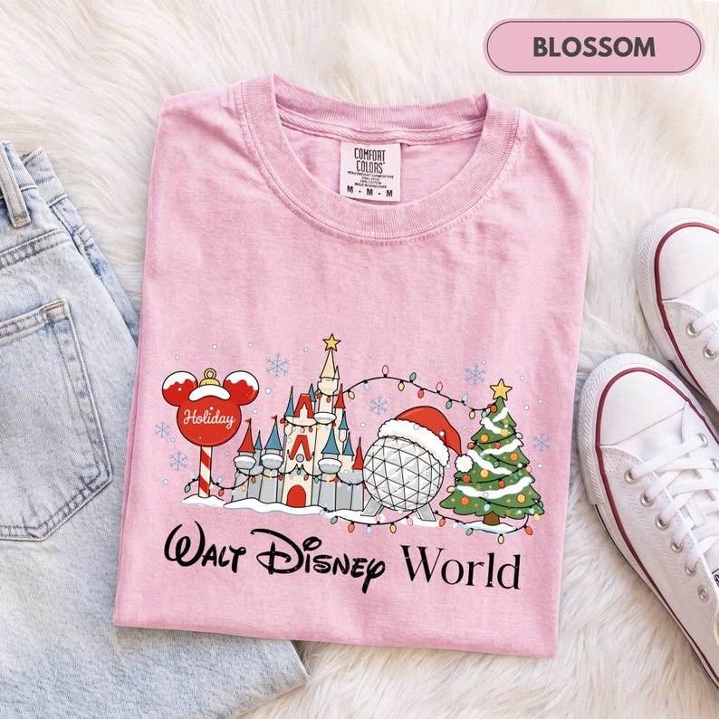 May include: A light pink short-sleeve t-shirt featuring a graphic of a holiday-themed castle, a geodesic sphere wearing a Santa hat, a Christmas tree, and a Mickey Mouse ornament. The text Walt Disney World is printed in black below.