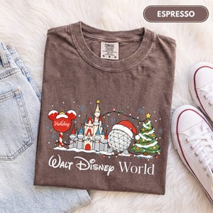 May include: An espresso brown t-shirt featuring a graphic of a castle, a geodesic sphere wearing a Santa hat, a Christmas tree, and a Mickey Mouse-shaped ornament. The text reads Walt Disney World and Holiday. The shirt has a crew neck and short sleeves.