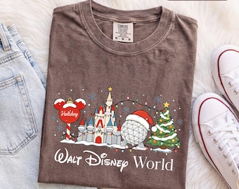 Könnte beinhalten: Ein espressobraunes T-Shirt mit einer Grafik, die ein Schloss, eine geodätische Kuppel mit Weihnachtsmütze, einen Weihnachtsbaum und einen Mickey-Mouse-förmigen Christbaumschmuck zeigt. Der Text lautet Walt Disney World und Holiday.