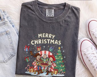 Könnte beinhalten: Ein anthrazitfarbenes T-Shirt mit einer Grafik von Woody, Buzz Lightyear und Jessie aus Toy Story, die zusammen einen Kuchen essen. Der Text Merry Christmas ist von bunten Geschenken, Zuckerstangen und einem Weihnachtsbaum umgeben.