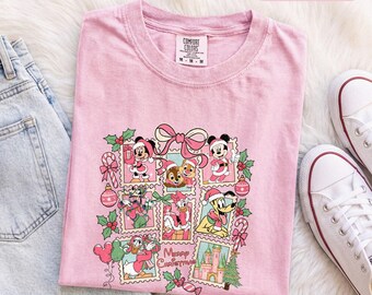 Könnte beinhalten: Ein hellrosa T-Shirt mit einem Grafikdruck von Disney-Figuren in Weihnachtskleidung, darunter Mickey, Minnie, Donald, Daisy, Pluto sowie Chip und Chap, angeordnet in Briefmarkenrahmen mit dem Text Merry Christmas.