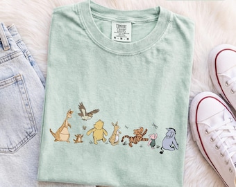 Könnte beinhalten: Ein hellgrünes Kurzarm-T-Shirt mit einem Grafikdruck der Winnie Puuh-Figuren, darunter Känga, Ruh, Eule, Puuh, Rabbit, Tigger, Ferkel und I-Aah. Das Shirt hat einen Rundhalsausschnitt und ein Comfort Colors-Markenetikett am Halsausschnitt.