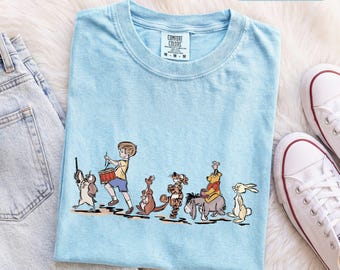 Könnte beinhalten: Ein hellblaues T-Shirt mit einer grafischen Illustration von Christopher Robin, der eine Trommel spielt, gefolgt von einer Reihe von Winnie Puuh-Figuren, darunter Roo, Tigger, I-Aah und Rabbit. Das Comfort Colors Etikett zeigt die Größe M.