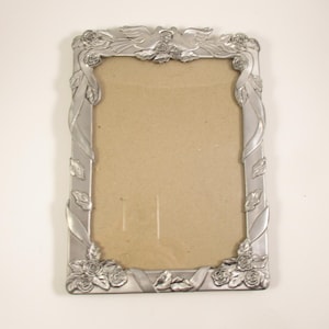 May include: A rectangular silver picture frame with floral and ribbon detailing. The frame has roses at the corners and top, with leaves and ribbons along the sides. The frame is empty, with a cardboard backing.