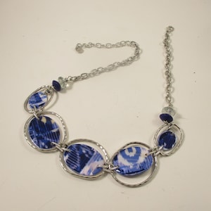 May include: A silver necklace with blue and white patterned oval and circular pendants. The pendants are linked by silver rings and chains. The necklace has a lobster clasp and is displayed on a white surface.