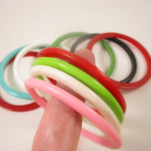 May include: A selection of colourful bangles in shades of red, pink, green, white, and blue. The round bracelets are stacked together, displaying a vibrant array of accessories. The bangles are made of a smooth, glossy material.