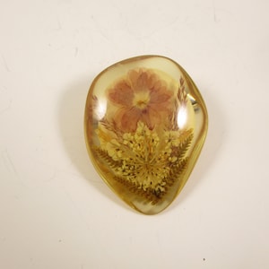 May include: A clear, teardrop-shaped resin pendant with a preserved flower and foliage. The flower is a muted reddish-brown, surrounded by yellow and green dried plants. The resin has a golden hue.