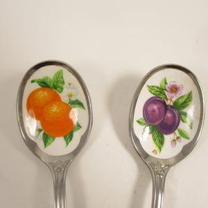 May include: Two decorative spoons with oval bowls. One spoon features a design of two oranges with green leaves and white flowers. The other spoon has a design of two purple plums with green leaves and pink flowers. The spoons have silver handles.