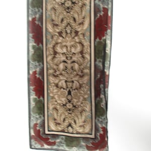 May include: A rectangular scarf featuring a central panel with a tan and brown botanical pattern on a black background, bordered by a light blue frame. The outer edges display a repeating pattern of large red and green floral motifs on a light blue base.