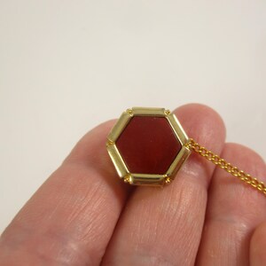 May include: A gold-coloured hexagonal pendant with a deep red centre stone. The pendant is attached to a delicate gold chain. The jewellery is held in a person's hand against a plain background.