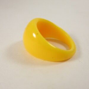 May include: A vibrant yellow ring with a smooth, glossy surface. The ring features a rounded, domed top and a thick band. The ring is a solid colour and appears to be made of plastic or resin.