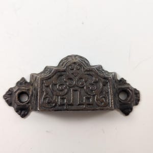 May include: A dark bronze drawer pull with an ornate, symmetrical design. The hardware features a central decorative element with scrolling patterns and two holes for mounting. The pull is likely made of metal and has a vintage aesthetic.