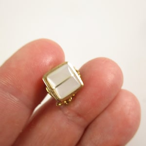 May include: A small, square-shaped vintage jewellery piece with a gold-coloured frame and a white, rectangular centre. The piece is held between fingers, showcasing its size and design. The jewellery has a classic, elegant aesthetic.