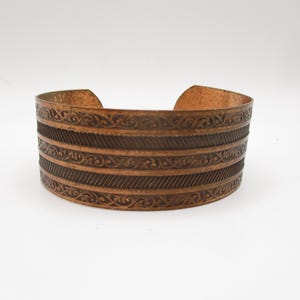 Peut inclure: Un large bracelet manchette en cuivre avec des motifs en relief complexes. Le bracelet présente des bandes alternées de volutes détaillées et de lignes texturées. Le design ouvert permet un port facile et met en valeur les tons métalliques chauds du cuivre.