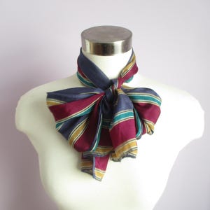 May include: A colourful striped neck scarf in navy, burgundy, teal, and gold is displayed on a white mannequin. The scarf is tied in a bow, showing the pattern. The scarf is made of a silky material.