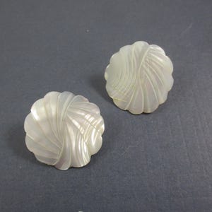 May include: A pair of white, shell-shaped earrings. Each earring has a swirling, textured design with a scalloped edge. The earrings are set against a grey background, showcasing their pearlescent sheen.
