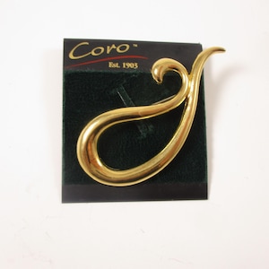 May include: A gold-coloured brooch with a fluid, abstract design, displayed on a dark green velvet card. The brooch has a smooth, polished surface and a distinctive shape. The card displays the brand name "Coro" and the text "Est. 1903".