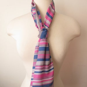 May include: A rectangular scarf featuring a repeating pattern of horizontal stripes in shades of hot pink, royal blue, light grey, and white. The scarf is tied in a simple knot at the centre, with the fabric ends hanging down in a straight vertical line.