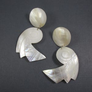 May include: Pair of iridescent white earrings. Each earring has a round top and a stylized, curved bottom with geometric and circular etched designs. The earrings are made of a pearlescent material.