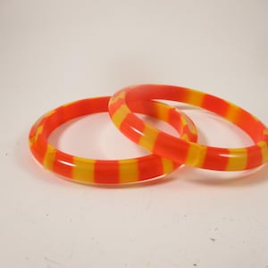 May include: Two orange and yellow striped bangles. The bracelets are made of a translucent material and are resting on a white surface. The bangles are a bright and colourful accessory.