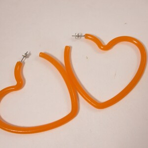 May include: A pair of orange heart-shaped hoop earrings. The earrings are made of a translucent orange material and have silver-colored posts. The earrings are displayed on a white surface.