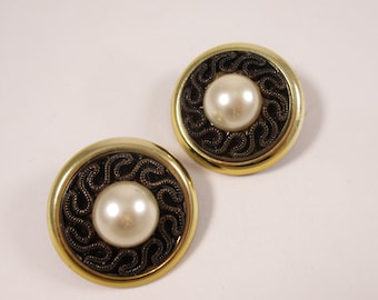 May include: Two round, gold-toned earrings with a central faux pearl. Each earring features a dark, swirling design around the pearl. The earrings are set against a white background, highlighting their design.