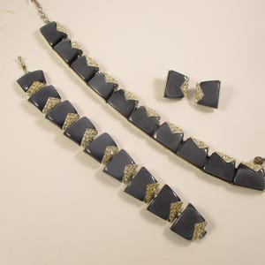 May include: A vintage jewellery set comprising a necklace, bracelet, and matching earrings. The set features a dark grey geometric design with silver-tone metal and sparkling accents. The bracelet and necklace are linked with a chain.