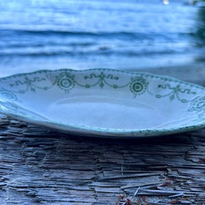May include: An oval-shaped ceramic dish with a cream-coloured base and a scalloped edge. The rim features a decorative green pattern consisting of circular motifs connected by thin lines and swags, creating a repeating border design.