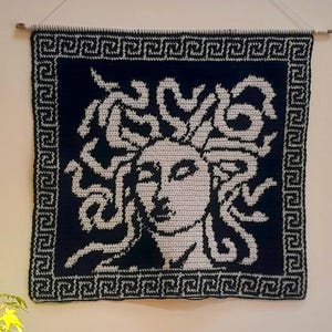 Peut inclure: Une tenture murale carrée en crochet noir et blanc représentant un portrait central de visage aux cheveux en forme de serpents, entouré d'une bordure géométrique à motif grec. La pièce textile est suspendue à une fine tige en bois.