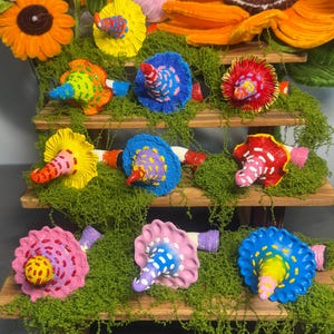 May include: A collection of eight handcrafted mushroom-shaped ornaments in various bright colors, including blue, pink, yellow, orange, and red. Each mushroom features a textured cap with contrasting painted dots and a stem, displayed on a wooden stand.