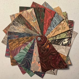 May include: A fan-shaped collection of rectangular paper swatches featuring various marbled patterns in diverse colors including deep red, forest green, slate blue, tan, and black. The swatches are secured together at one end by a silver metal screw.