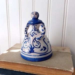 May include: A ceramic bell shaped like a stylized figure with a white base and blue hand-painted details. The figure features a rounded head, a wide skirt, and arms curved inward. The surface is decorated with blue floral and abstract patterns.