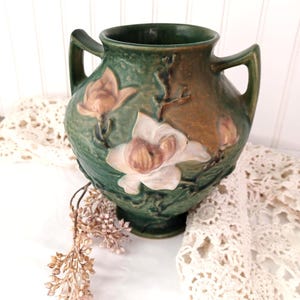 Puede incluir: Un jarrón de cerámica verde oscuro con dos asas, que presenta diseños florales en relieve en tonos blanco y rosa. La superficie tiene un acabado mate y texturizado con acentos marrones. El jarrón tiene un cuerpo ancho y redondeado.