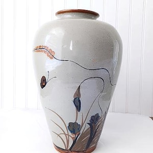 May include: A light gray ceramic vase with a speckled finish, featuring a hand-painted design of dark blue flowers, brown grasses, and a small butterfly. The vase has a smooth, rounded body, a narrow neck, and a brown glazed rim and base.