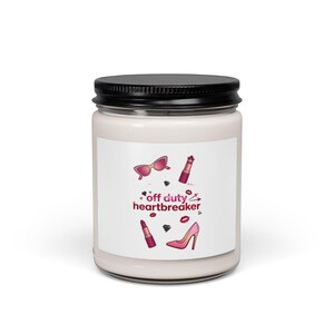 May include: A white candle in a clear glass jar with a black lid. The label features the text "off duty heartbreaker" in red, surrounded by illustrations of pink sunglasses, lipsticks, diamonds, lip prints, and a high-heeled shoe.