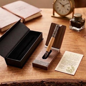 May include: A wooden pen with a metallic cap featuring intricate engravings, displayed in an open dark wood case. The pen has a light-colored wood barrel and a dark metal tip. A black gift box, a certificate of authenticity, and an ink bottle are nearby.