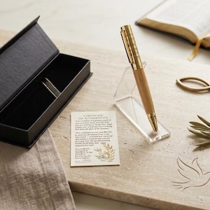 May include: A gold and light brown wooden pen rests on a clear acrylic stand next to an open black rectangular gift box. A certificate of authenticity card is placed nearby, featuring text and a small illustration of an olive branch.