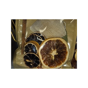 May include: A collection of dried citrus slices including dark brown and golden orange segments, accompanied by a single brown cinnamon stick and a small beige burlap sachet, all contained within a clear plastic packaging pouch.