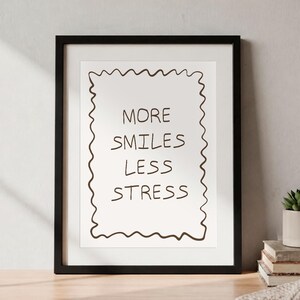 Pode incluir: Uma impressão artística emoldurada com fundo branco e uma borda ondulada desenhada à mão em castanho escuro. O texto "MORE SMILES LESS STRESS" está escrito no centro com um tipo de letra simples, manuscrito e castanho escuro.
