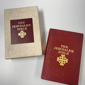 Puede incluir: Un ejemplar de la Biblia de Jerusalén encuadernado en cuero rojo con letras doradas y un emblema de cruz dorada, acompañado de su estuche protector color crema a juego con un panel rectangular rojo oscuro con el mismo título y cruz.